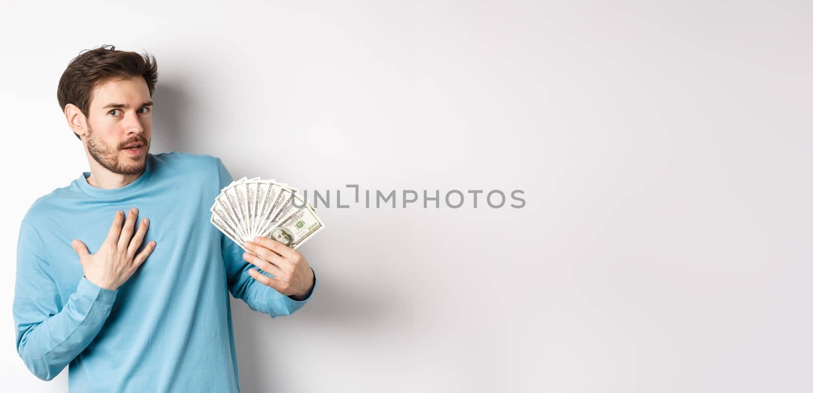 Surprised man holding money, looking amazed at camera, touching chest and gasping, standing in blue sweatshirt over white background by Benzoix