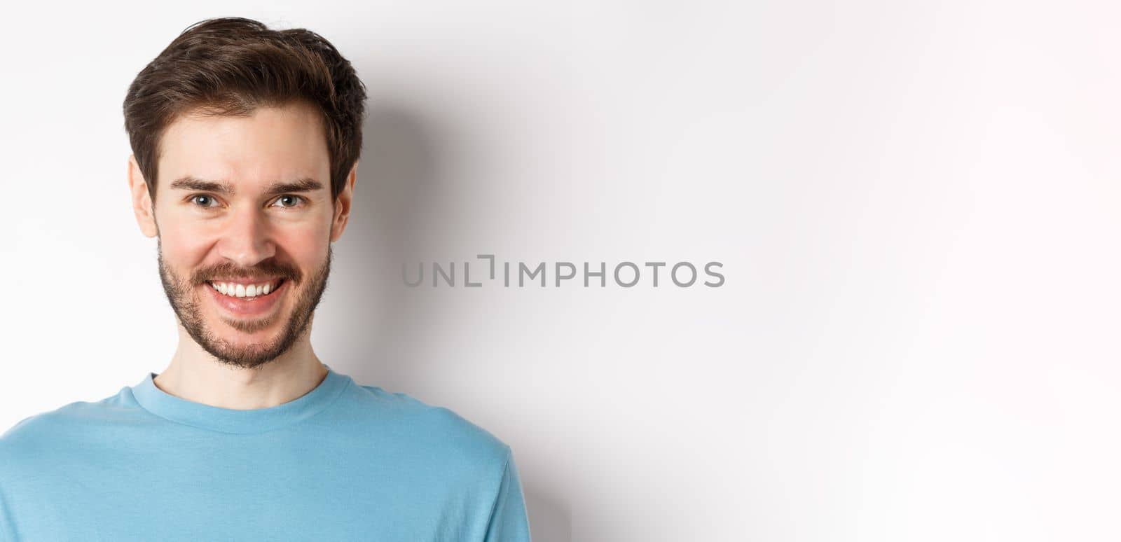 Close-up of handsome caucasian man smiling with white teeth, looking confident at camera, standing in blue shirt on white background.