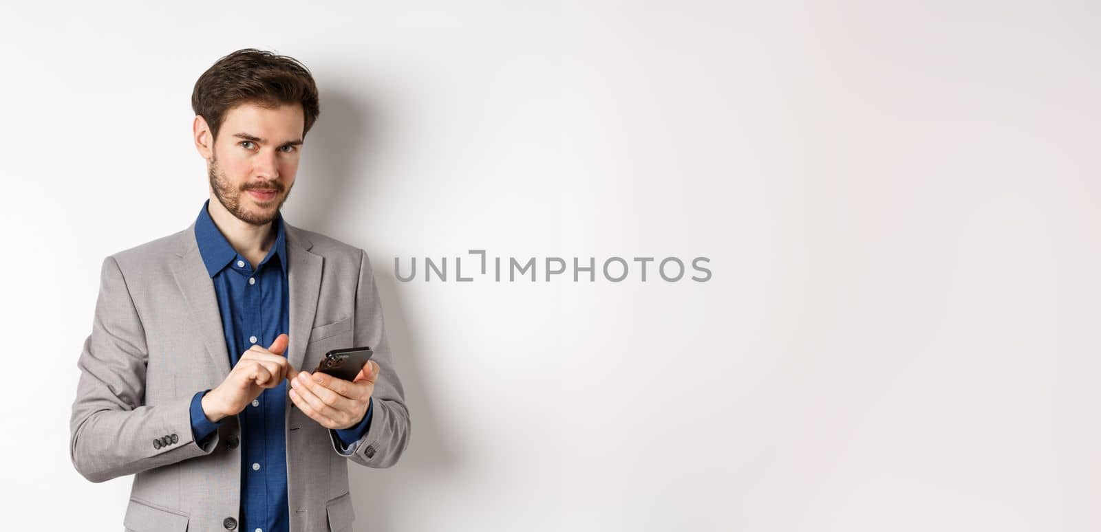 Handsome bearded male model in suit using mobile phone, smiling pleased at camera, white background.