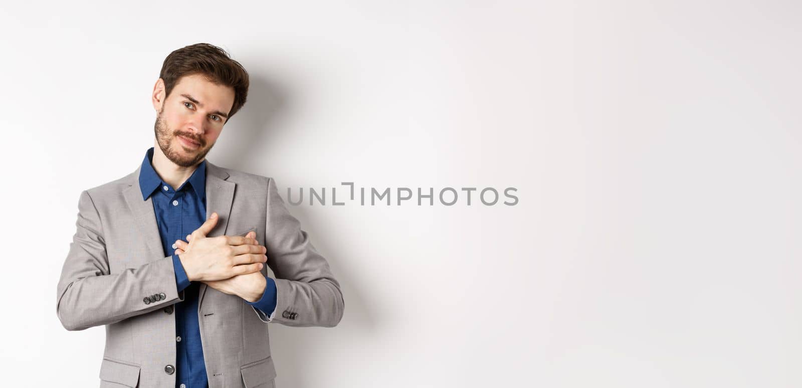 Romantic handsome guy with beard holding hands on heart and smiling, thanking you, feeling nostalgic, having tender memories in heart, white background.