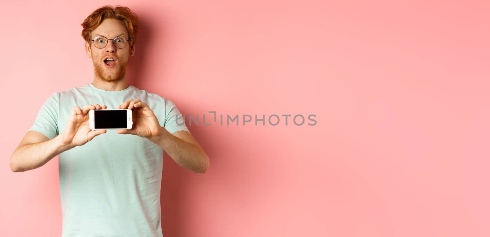 Amazed redhead man gasping and staring with awe at camera, showing blank smartphone screen horizontally, standing over pink background by Benzoix