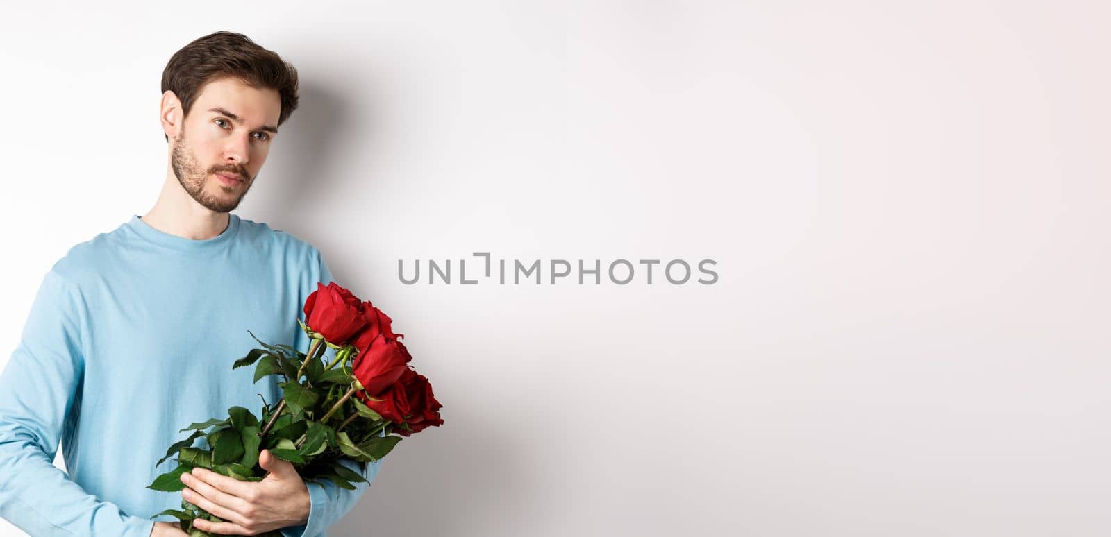 Handsome young man holding beautiful red roses for his lover on Valentines day, looking pensive, standing over white background.