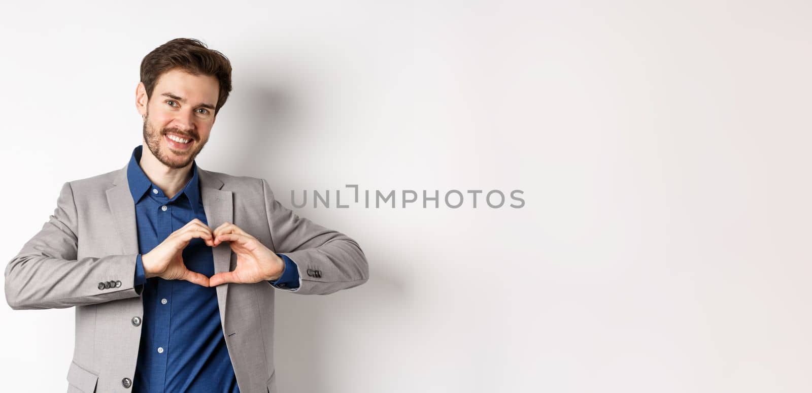 Romantic man in suit showing heart sign and smiling, love his girlfriend, standing on white background, making confession to lover.