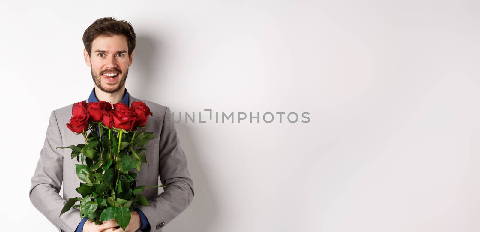 Excited handsome man in suit holding bouquet of roses for romantic date with lover, standing happy on Valentines day, white background by Benzoix
