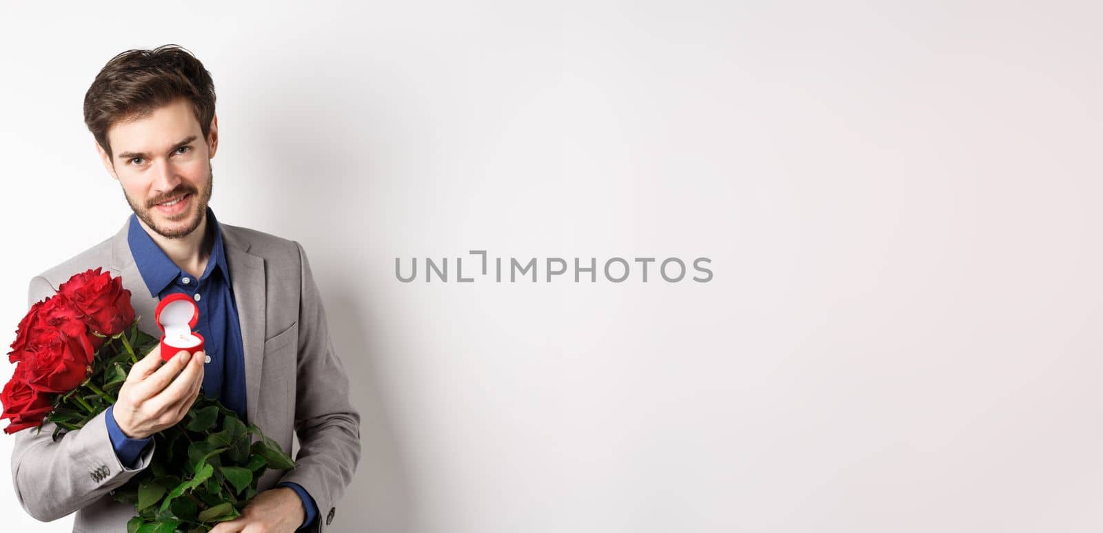 Romantic man with boquet of red roses asking to marry him, holding engagement ring and looking confident at camera, standing in suit over white background by Benzoix
