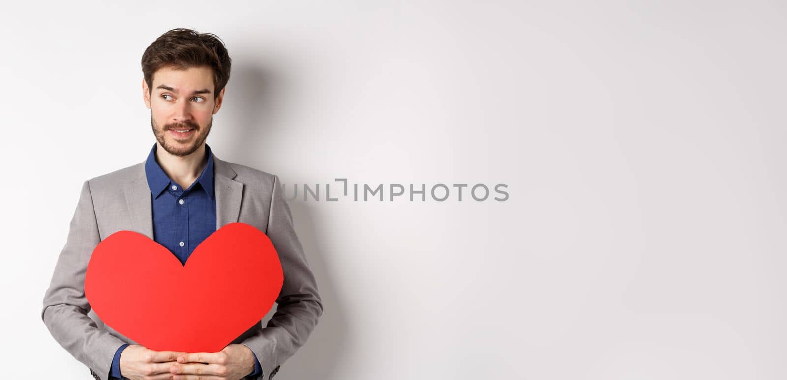 Attractive man in suit looking left and smiling, holding red heart cutout, prepare valentines day surprise for lover, standing over white background.