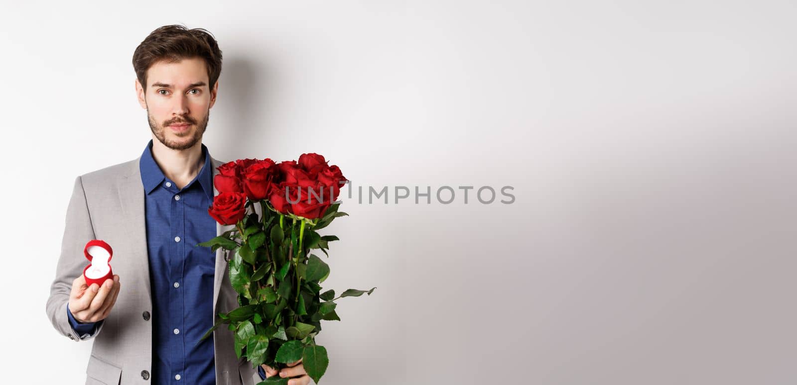 Handsome man in suit going to make a proposal, standing with red roses flowers and engagement rin in box, making romantic surprise, white background by Benzoix