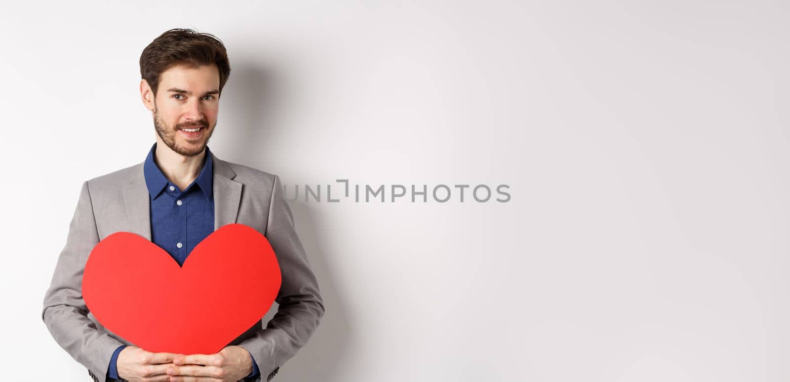 Handsome bearded man wishing happy valentines day, holding red heart cutout and smiling, going on romantic date in fancy suit, standing over white background.