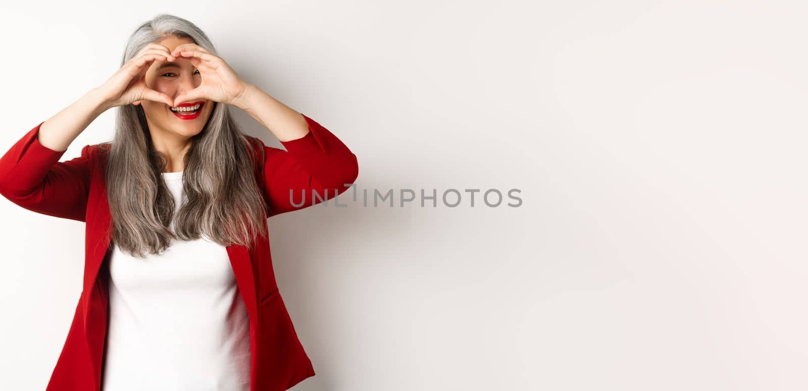 Lovely asian middle-aged woman with grey hair, wearing red blazer, showing heart sign and peeking throught it, I love you gesture, standing over white background by Benzoix