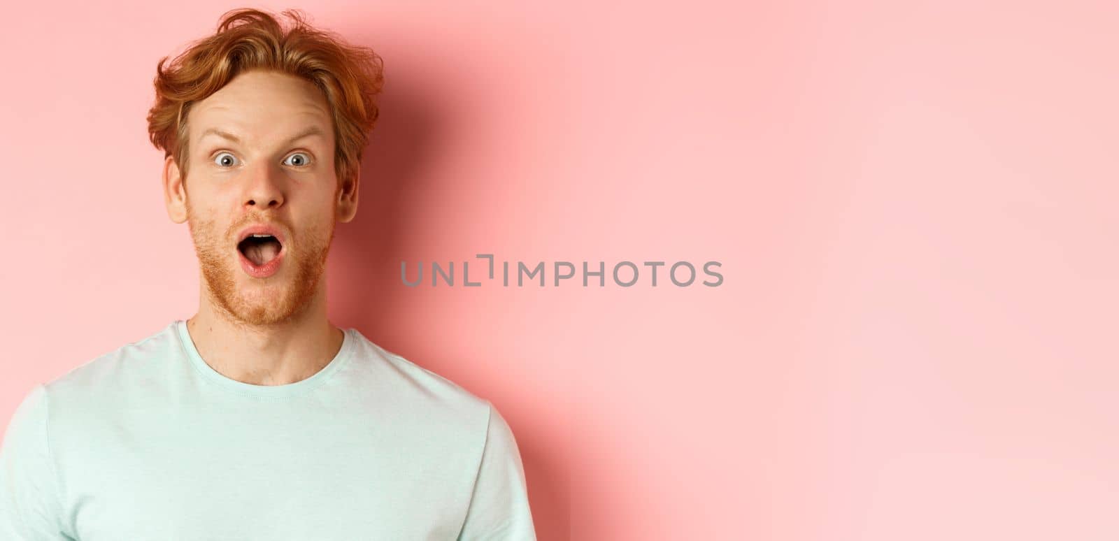 Face of surprised redhead man reacting to cool promo offer, raising eyebrows and gasping, staring in awe at camera, standing over pink background by Benzoix