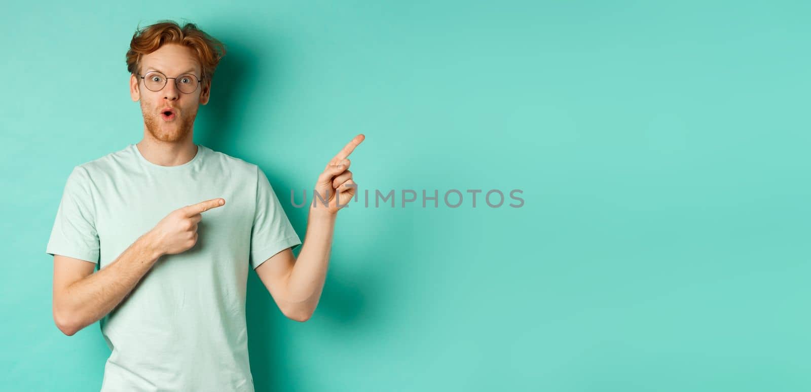 Surprised redhead guy in glasses and t-shirt checking out special deal, pointing at upper right corner promo and gasping in awe, showing banner, standing over turquoise background.