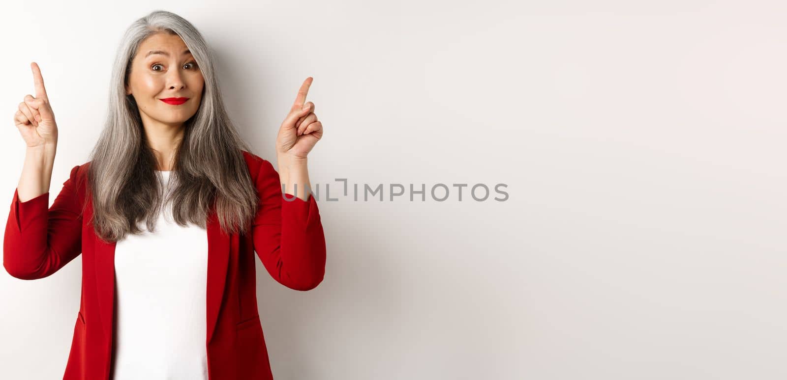 Elegant asian senior lady in red blazer and makeup, pointing fingers up and smiling, showing advertisement, white background.