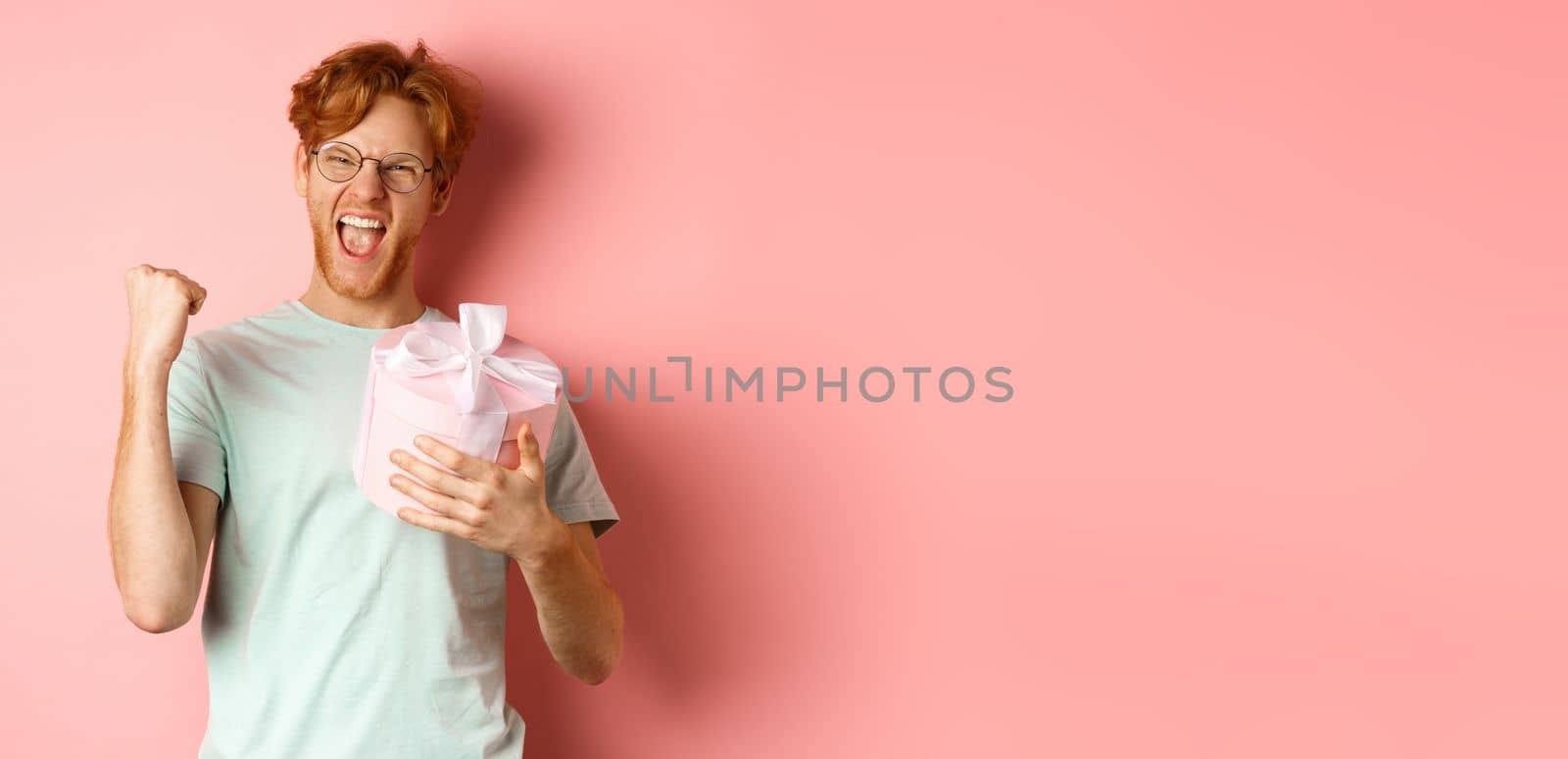 Valentines day and romance concept. Cheerful redhead guy making fist pump and scream yes with joy, holding gift box, smiling, standing over pink background by Benzoix
