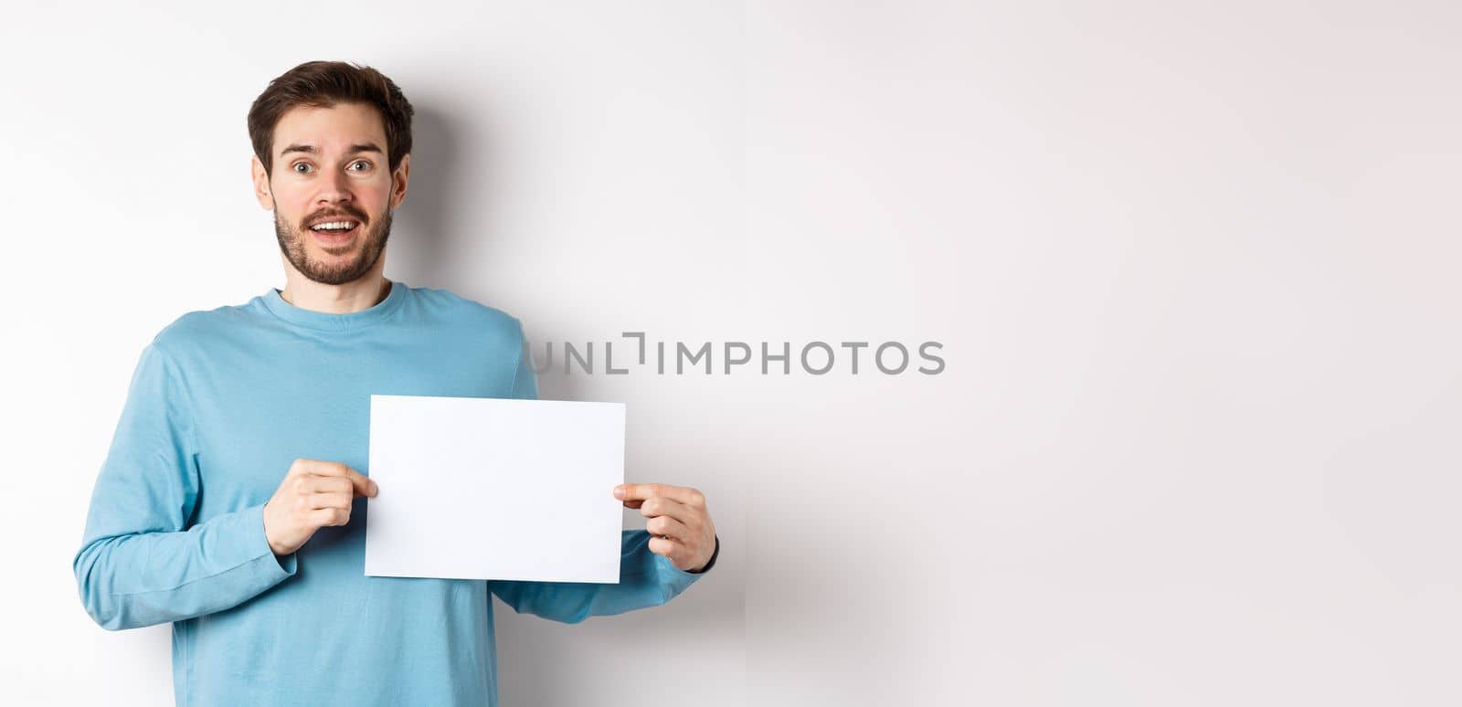 Romantic man looking with hopeful face at camera, showing sign card, blank piece of paper for banner or logo, standing over white background.
