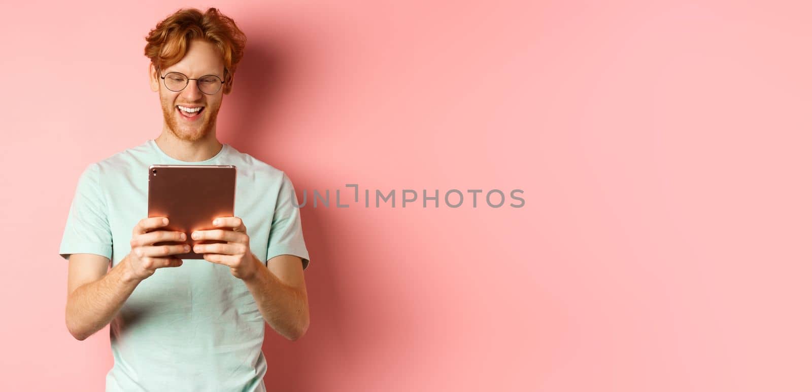 Happy guy with red hair and beard using digital tablet, reading screen and smiling, standing over pink background.