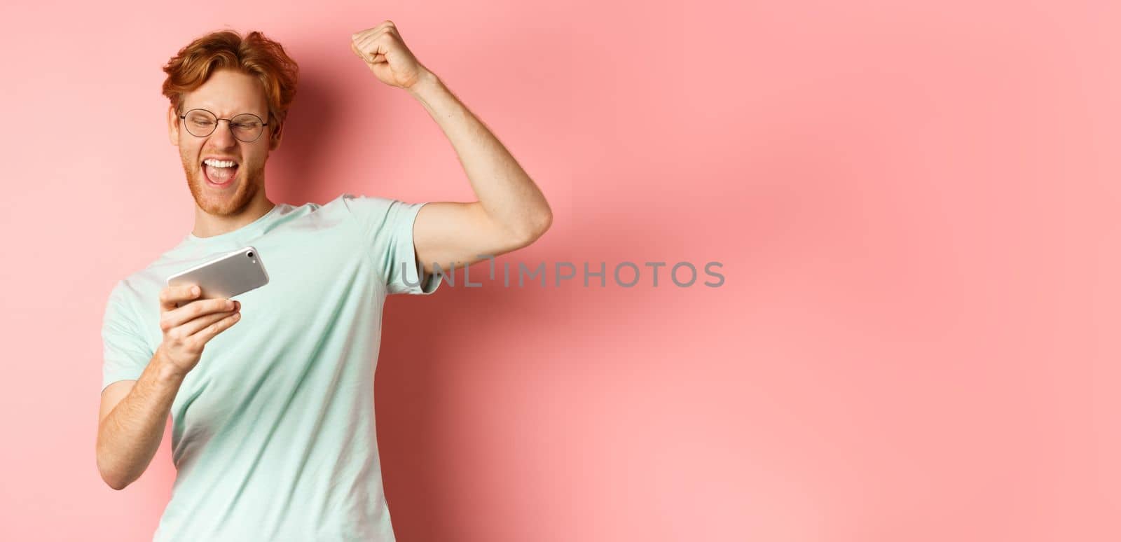 Happy redhead man winning in mobile video game, raising hand up and shouting yes with joy, celebrating victory, looking at smartphone, standing over pink background.