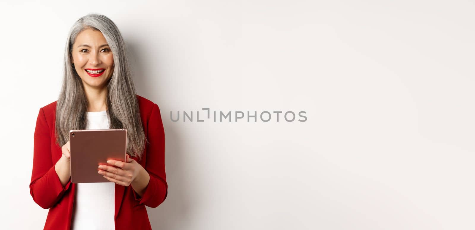 Business. Senior female entrepreneur working on digital tablet and smiling happy at camera, wearing red blazer and makeup, standing over white background.