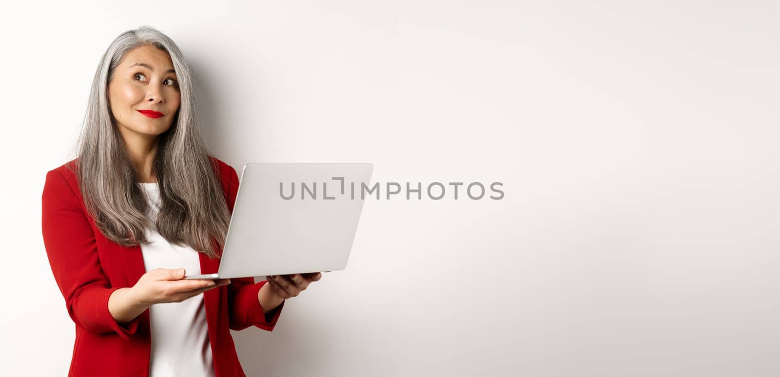 Business. Successful asian businesswoman in red blazer imaging something, working on laptop and looking upper left corner with dreamy smile, standing over white background by Benzoix