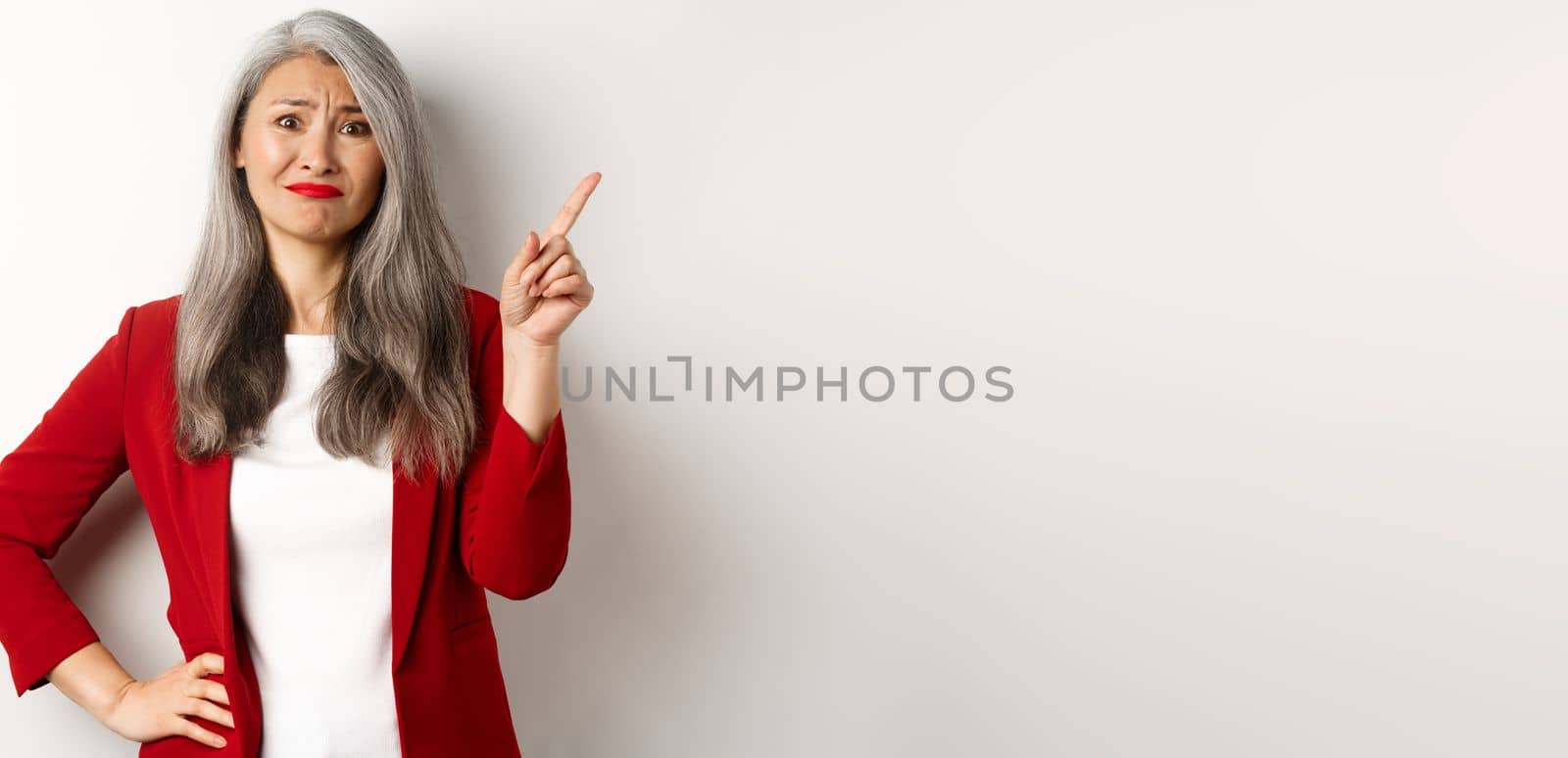 Disappointed and skeptical asian female entrepreneur pointing finger upper right corner, grimacing and frowning upset, showing bad promo, white background.