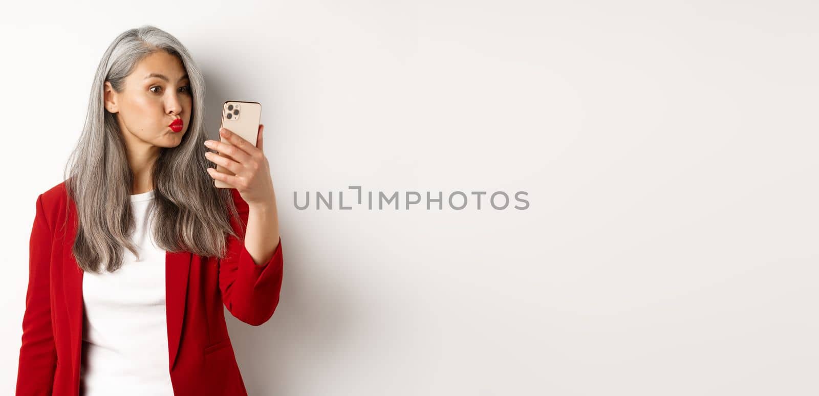 Silly and funny asian senior woman pouting, taking selfie on smartphone, standing over white background.