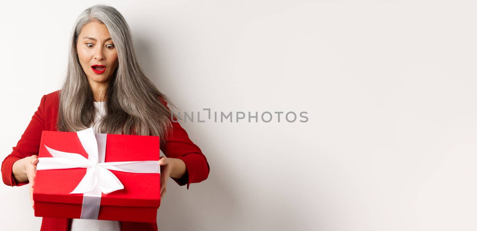 Surprised asian senior woman receiving present on mother day, holding red box with gift and looking amazed, white background by Benzoix