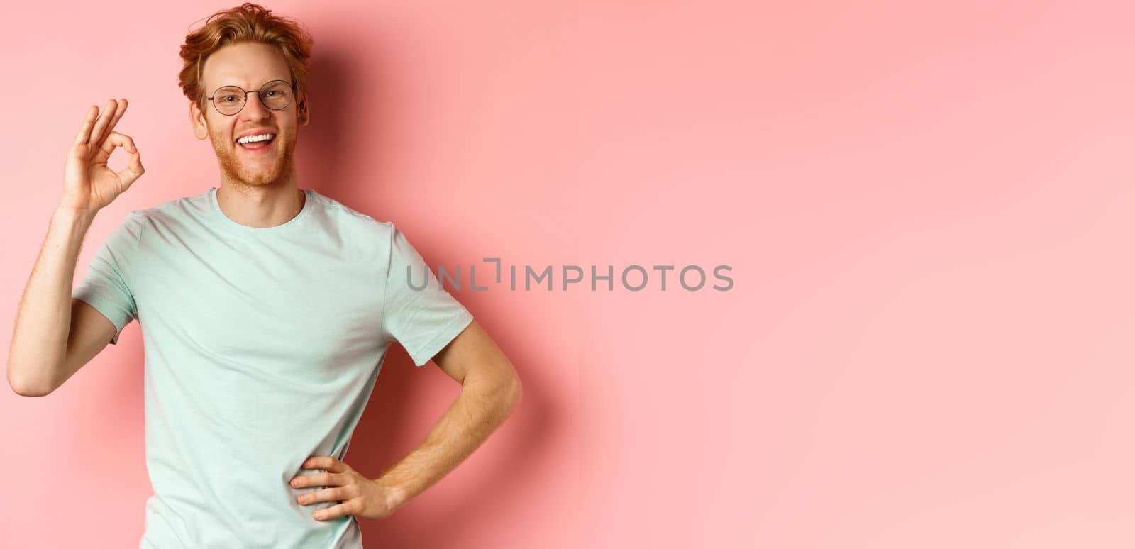 Cheerful guy with red hair and beard, wearing glasses, showing OK sign in approval and saying yes, smiling satisfied, standing over pink background.