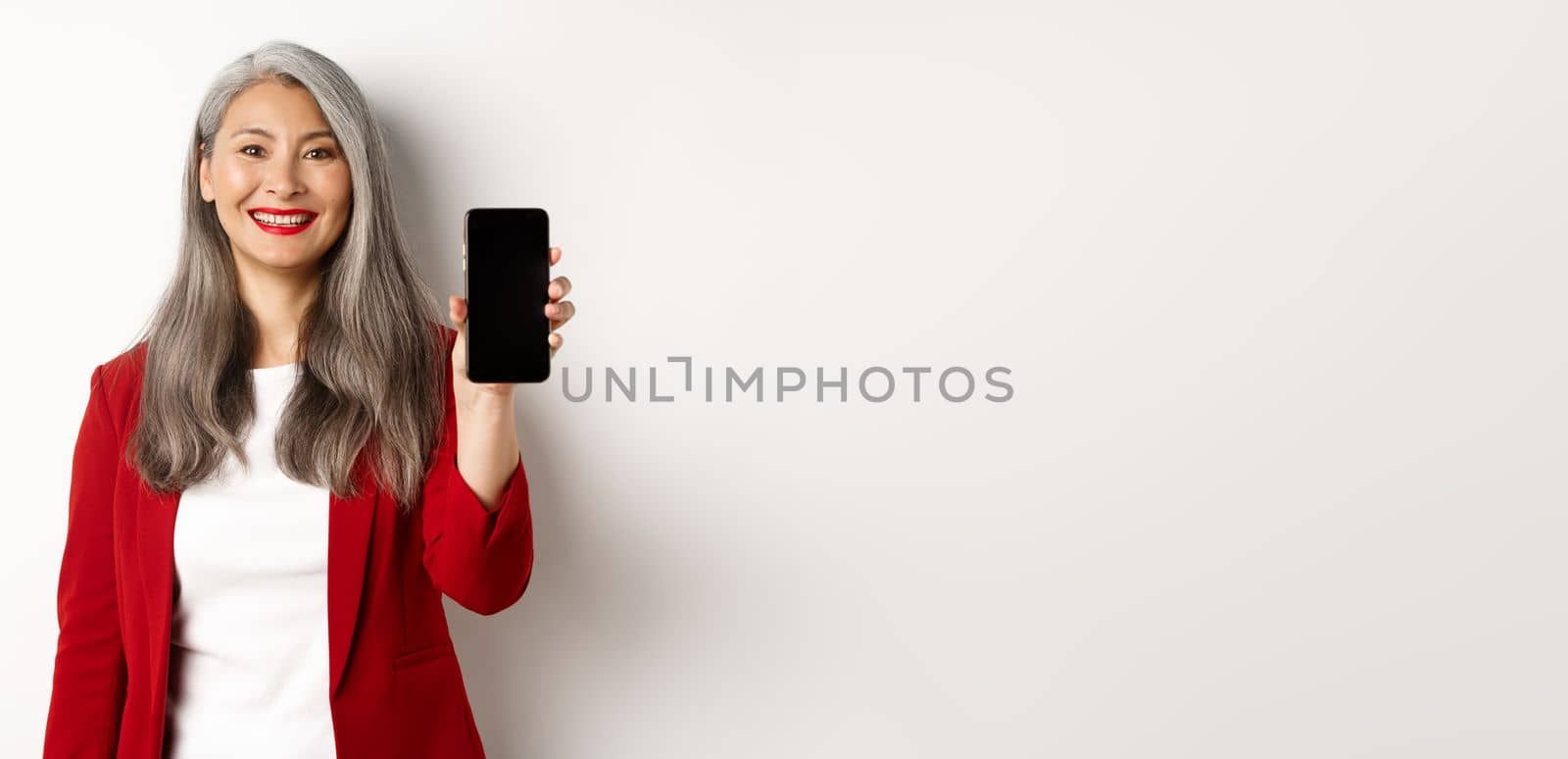 Elderly businesswoman in red blazer, showing blank smartphone screen and smiling, demonstrate mobile app, white background by Benzoix