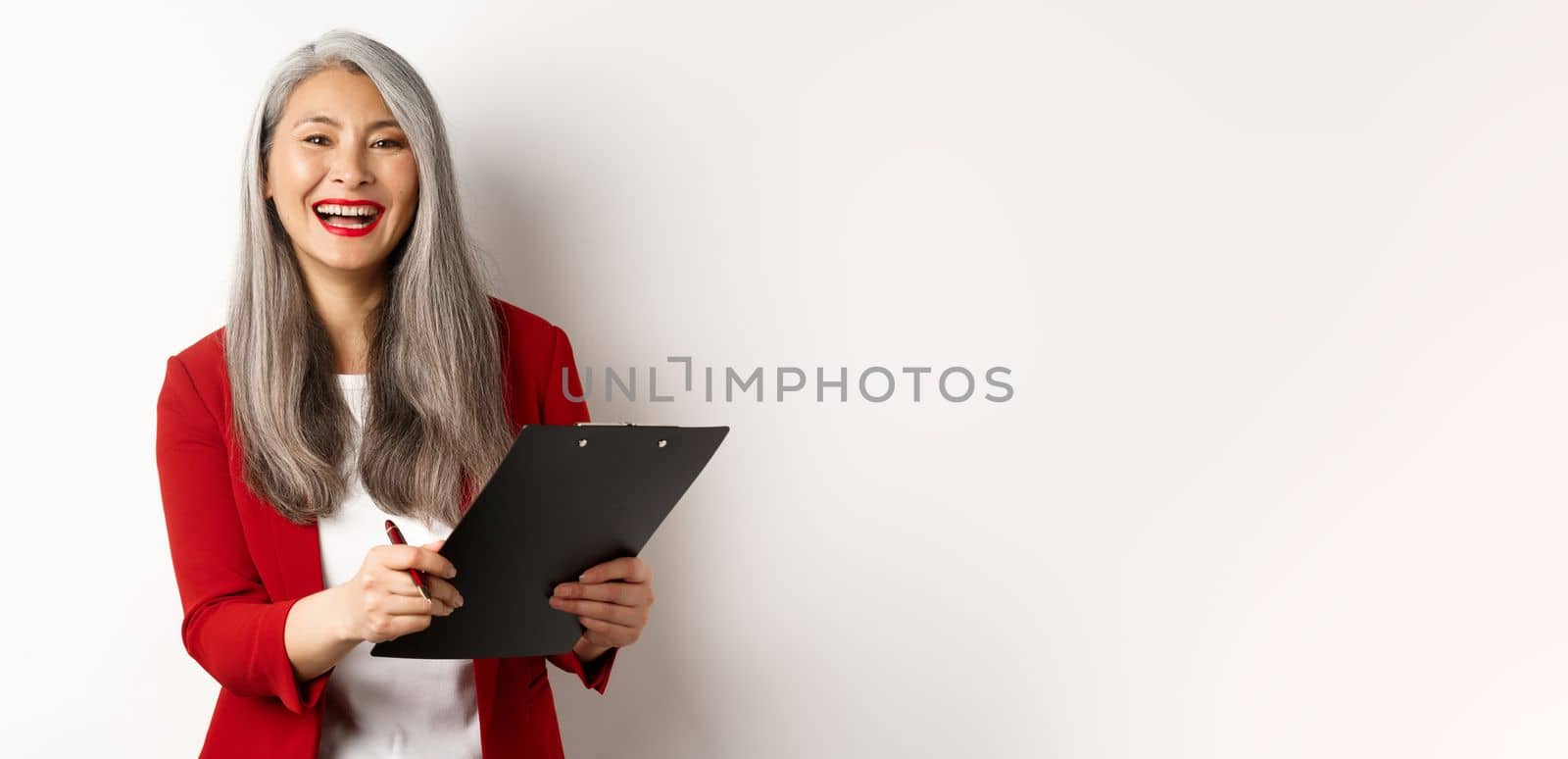 Happy asian office employee, woman in red blazer, working and laughing, holding clipboard with documents, standing over white background.