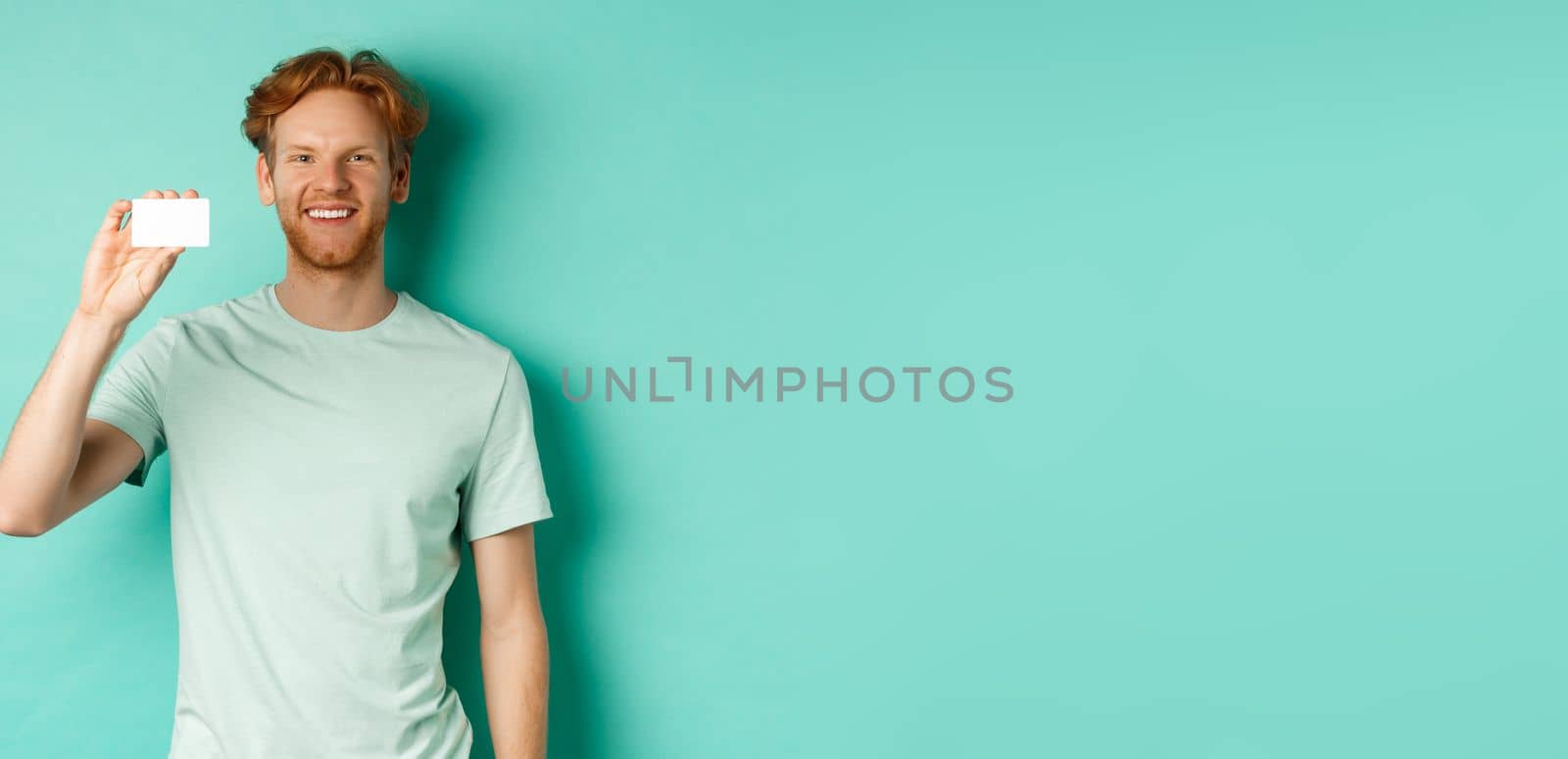 Shopping concept. Handsome redhead man in t-shirt showing plastic credit card and smiling, standing over turquoise background by Benzoix