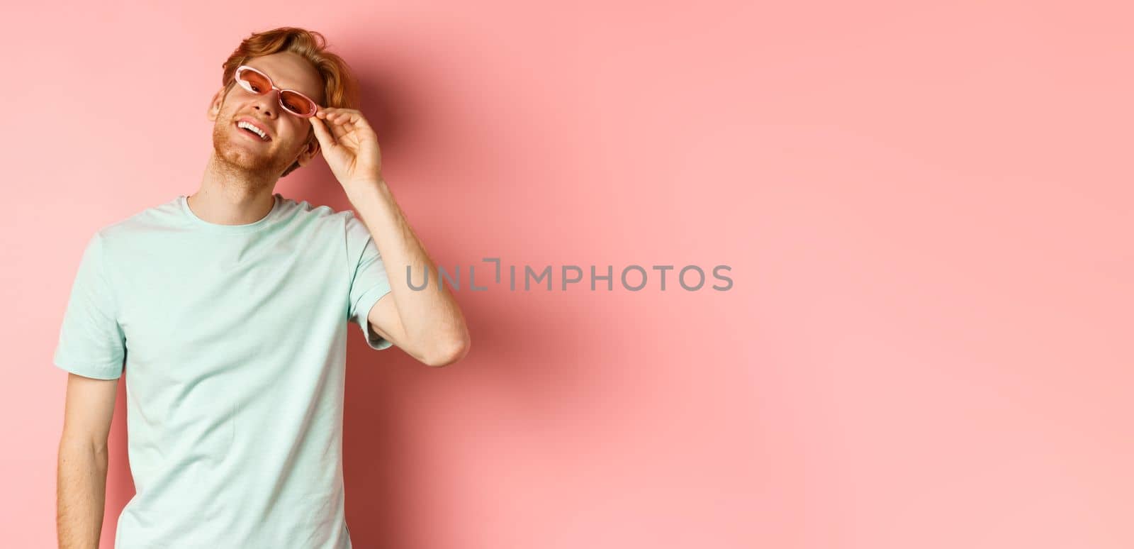 Tourism and vacation concept. Happy young man with red hair relaxing, wearing sunglasses and smiling carefree, standing over pink background.