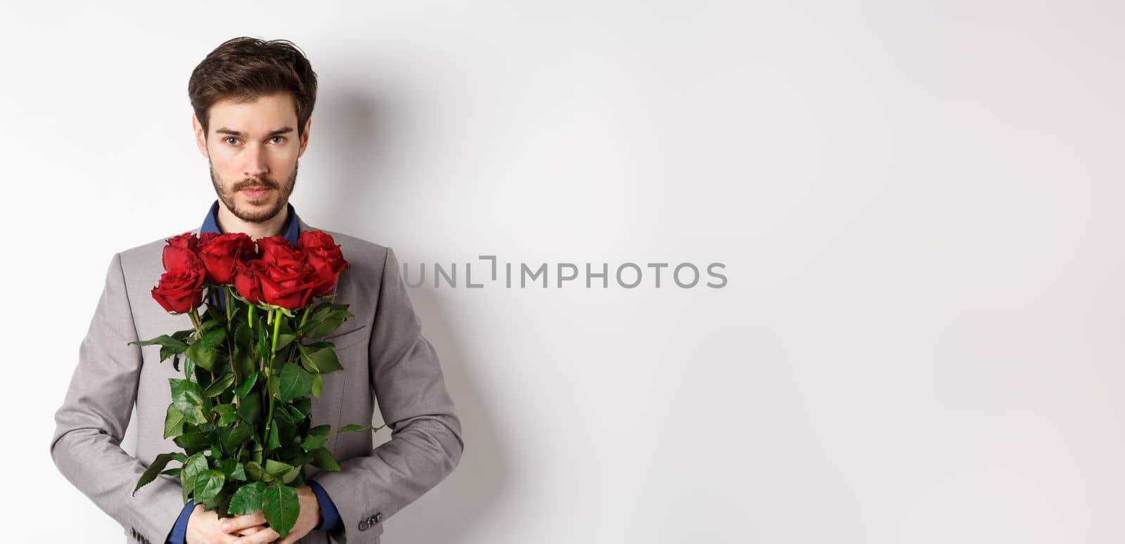 Valentines day and love concept. Confident and handsome man in suit holding bouquet of roses, looking at camera, standing over white background.