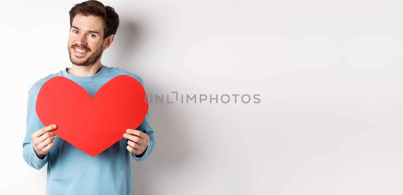 Handsome european man in sweater saying I love you, boyfriend standing with valentines day red heart, posing over white background by Benzoix