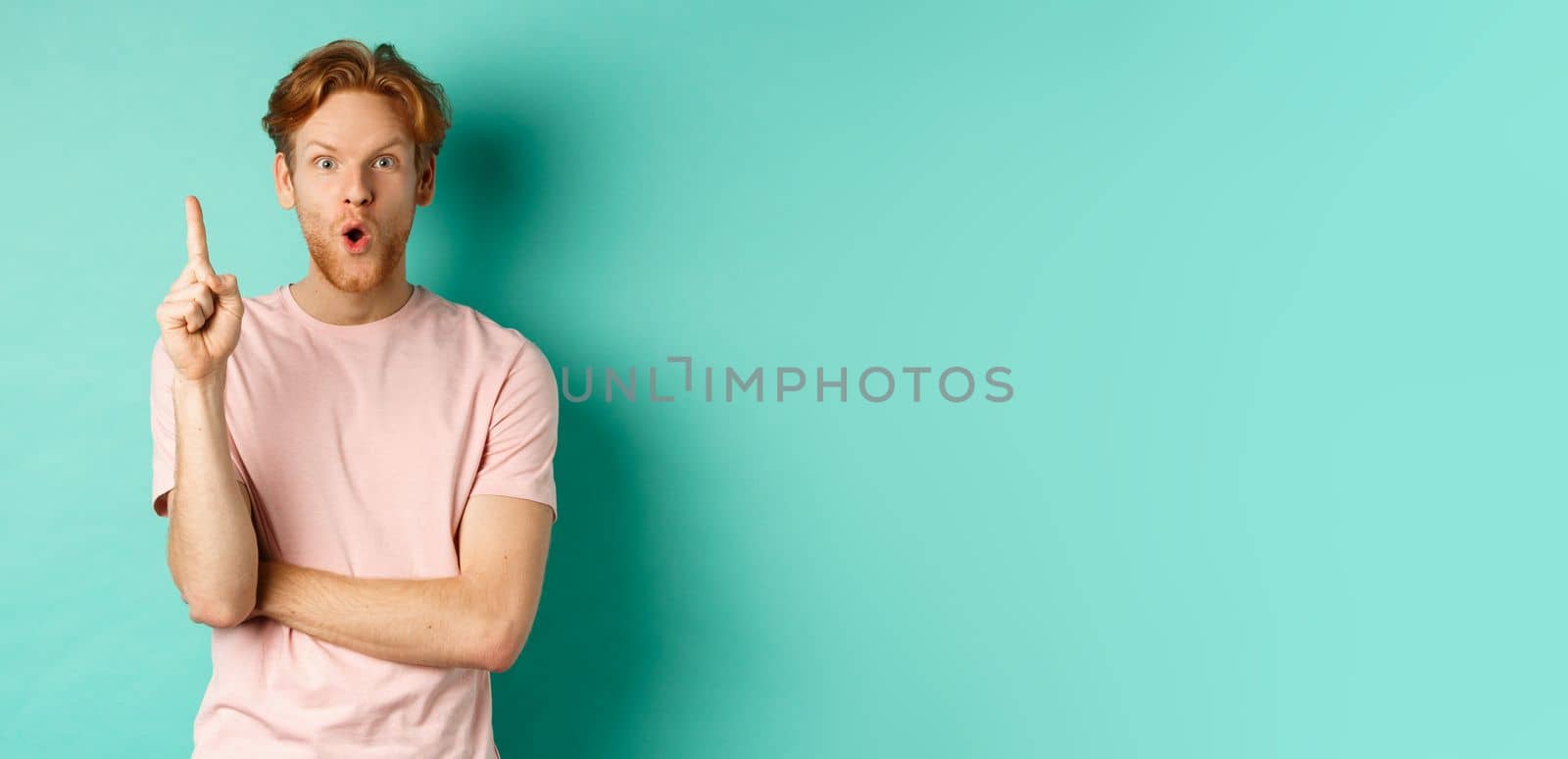 Pensive redhead man in t-shirt raising index finger, gasping as pitching at idea, saying suggestion, standing over turquoise background. Copy space