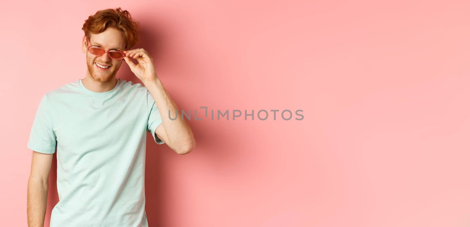 Tourism and vacation concept. Relaxed young man with red hair, standing in sunglasses and t-shirt and smiling pleased, standing over pink background by Benzoix