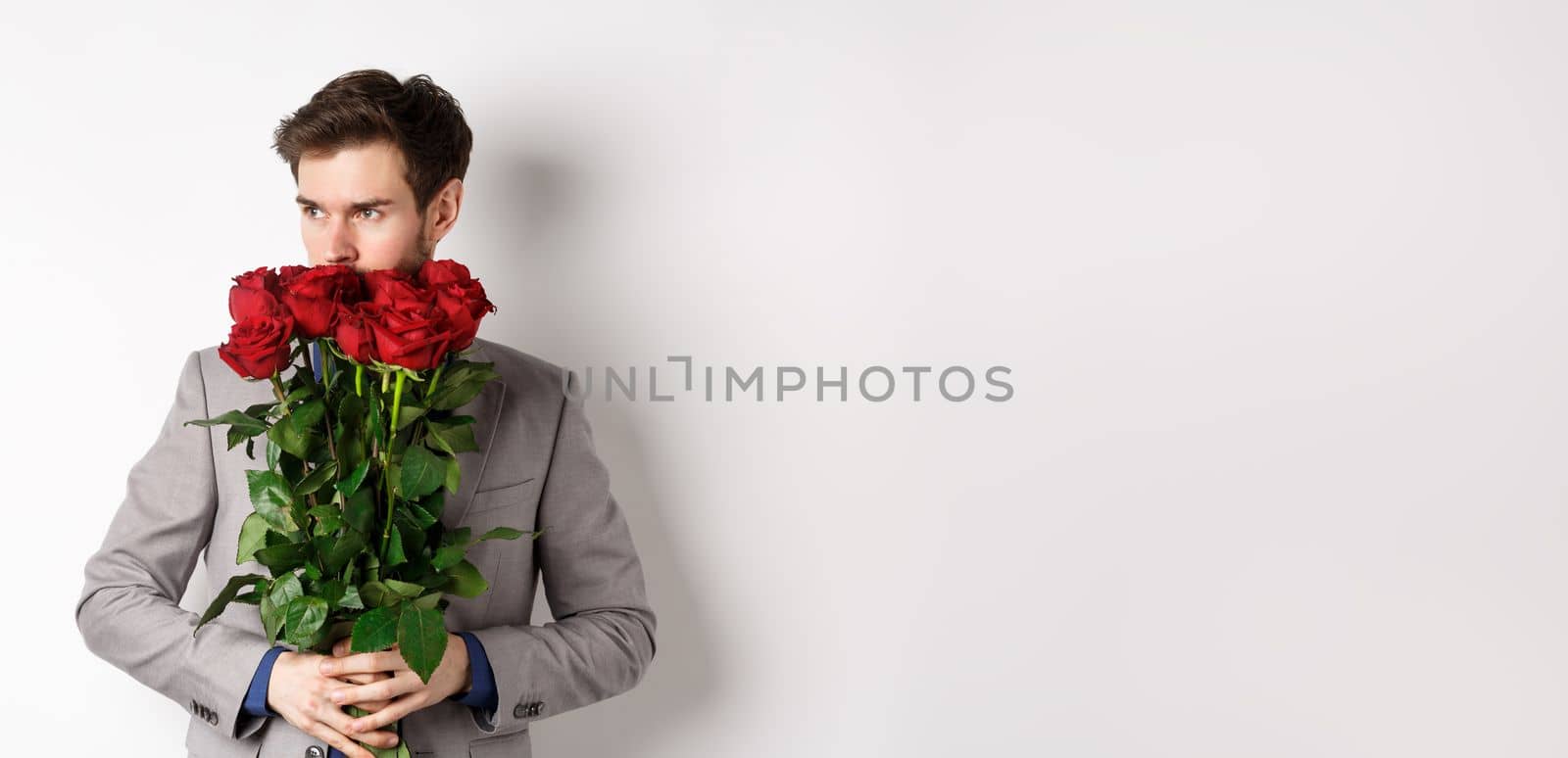 Romantic man in suit smell bouquet of roses and looking pensive, standing over white background. Concept of valentines day.