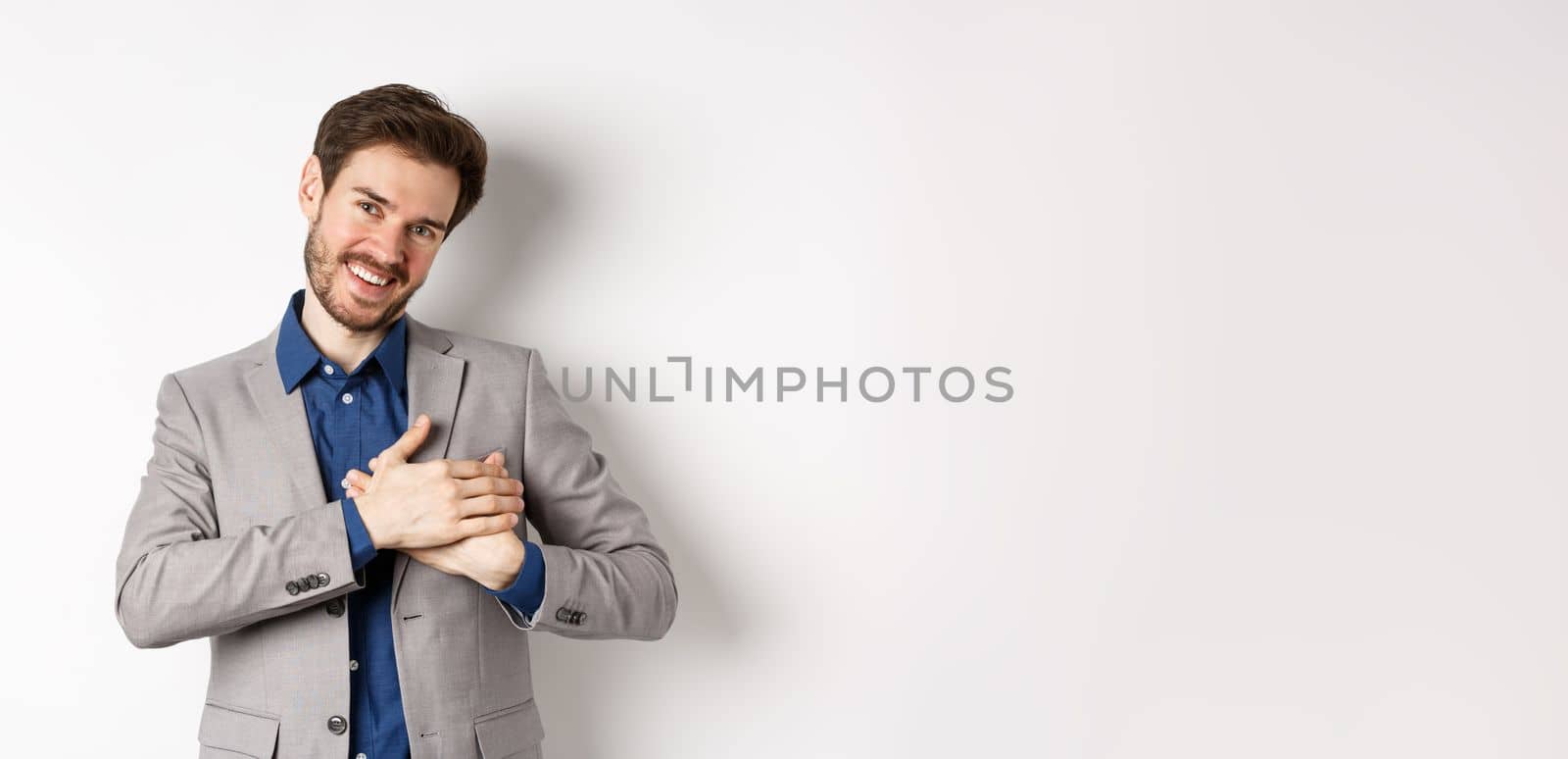 Happy man in suit holding hands on heart, feeling touched or grateful, having tender memories in soul, standing against white background.