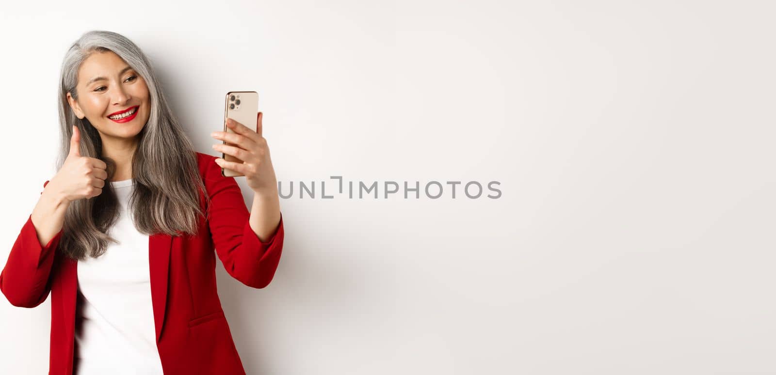 Successful asian businesswoman in red blazer, taking selfie on smartphone with thumb-up, showing approval, standing over white background.