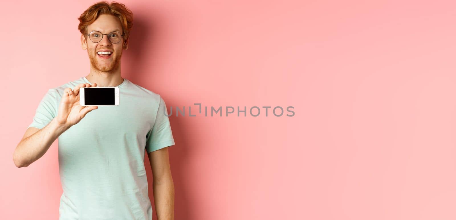 Happy young man with red hair showing smartphone screen, holding phone horizontally and smiling amazed, standing over pink background by Benzoix