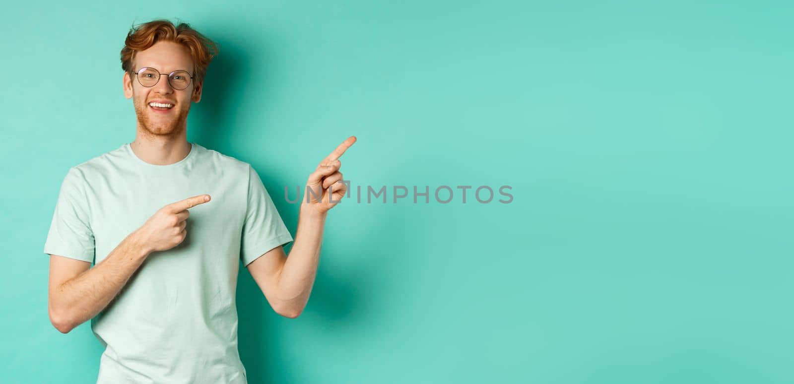 Handsome caucasian man with ginger hair, wearing glasses and t-shirt, pointing fingers right and smiling joyful, showing advertisement, standing over turquoise background.