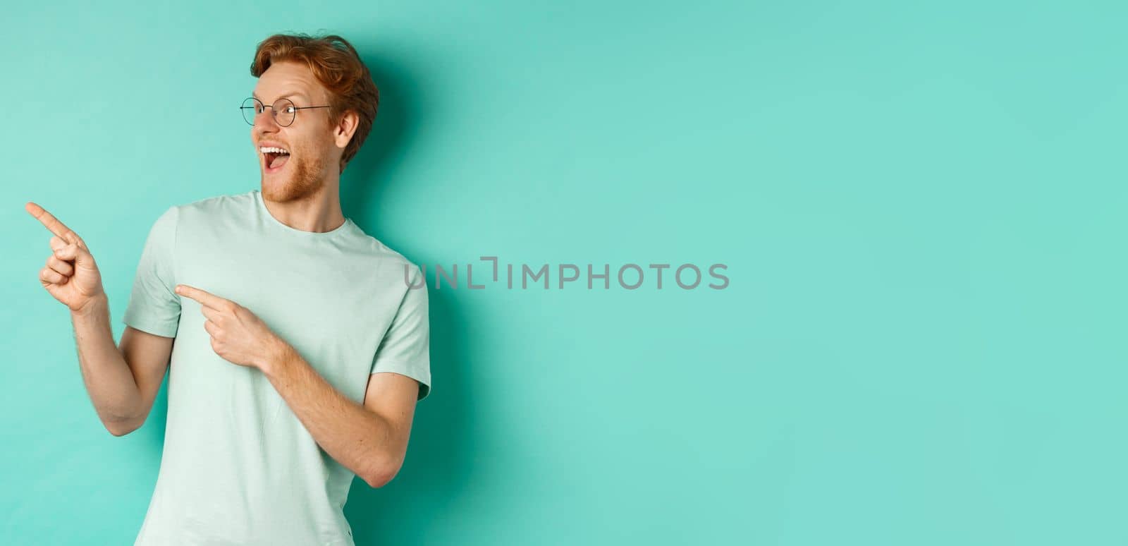 Excited young man with red hair, wearing glasses and t-shirt, pointing and looking left at awesome promotion, smiling happy at banner, turquoise background.