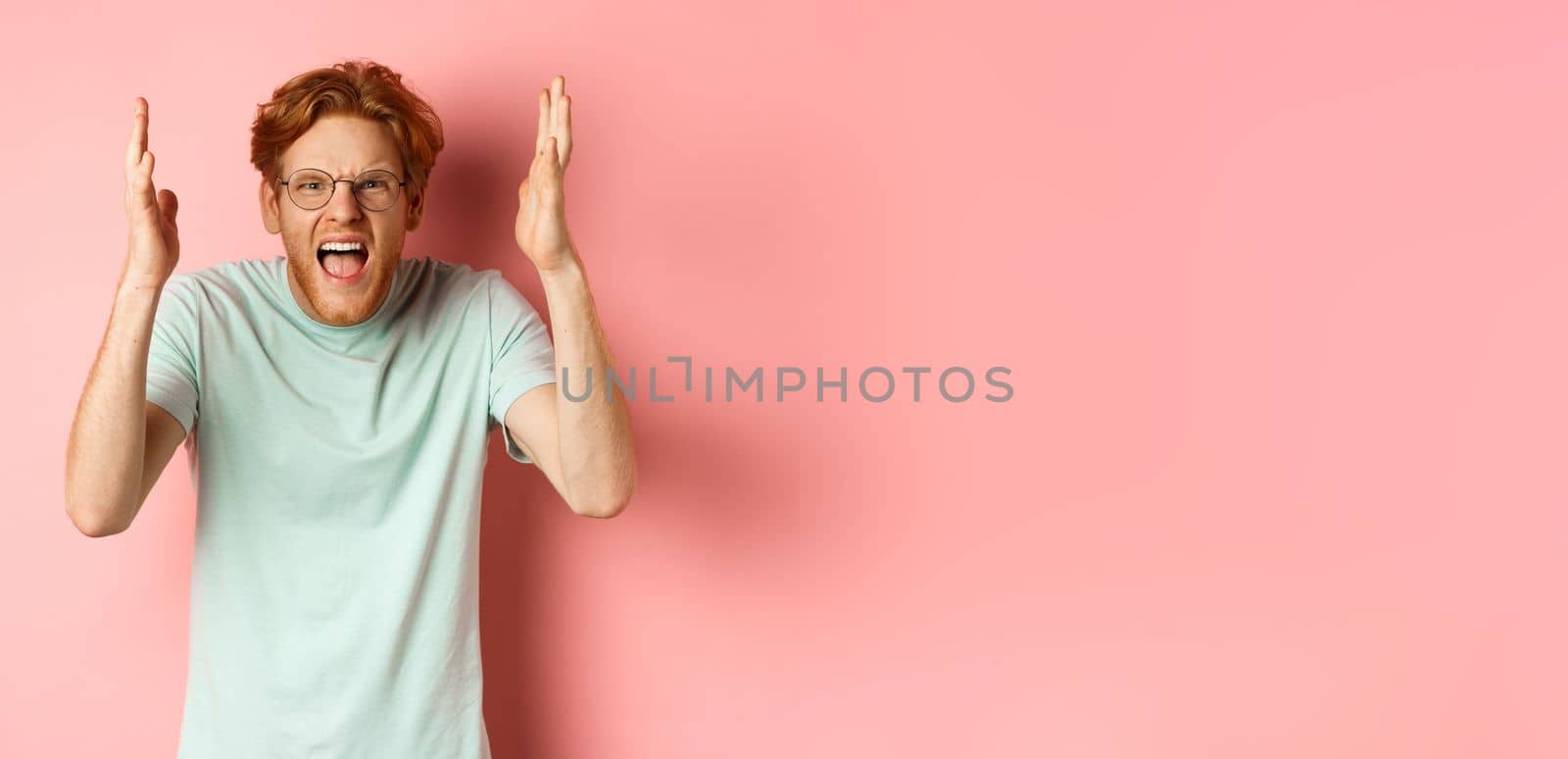 Angry redhead guy in glasses shouting, frowning and shaking hands with frustrated and outraged face, standing over pink background.
