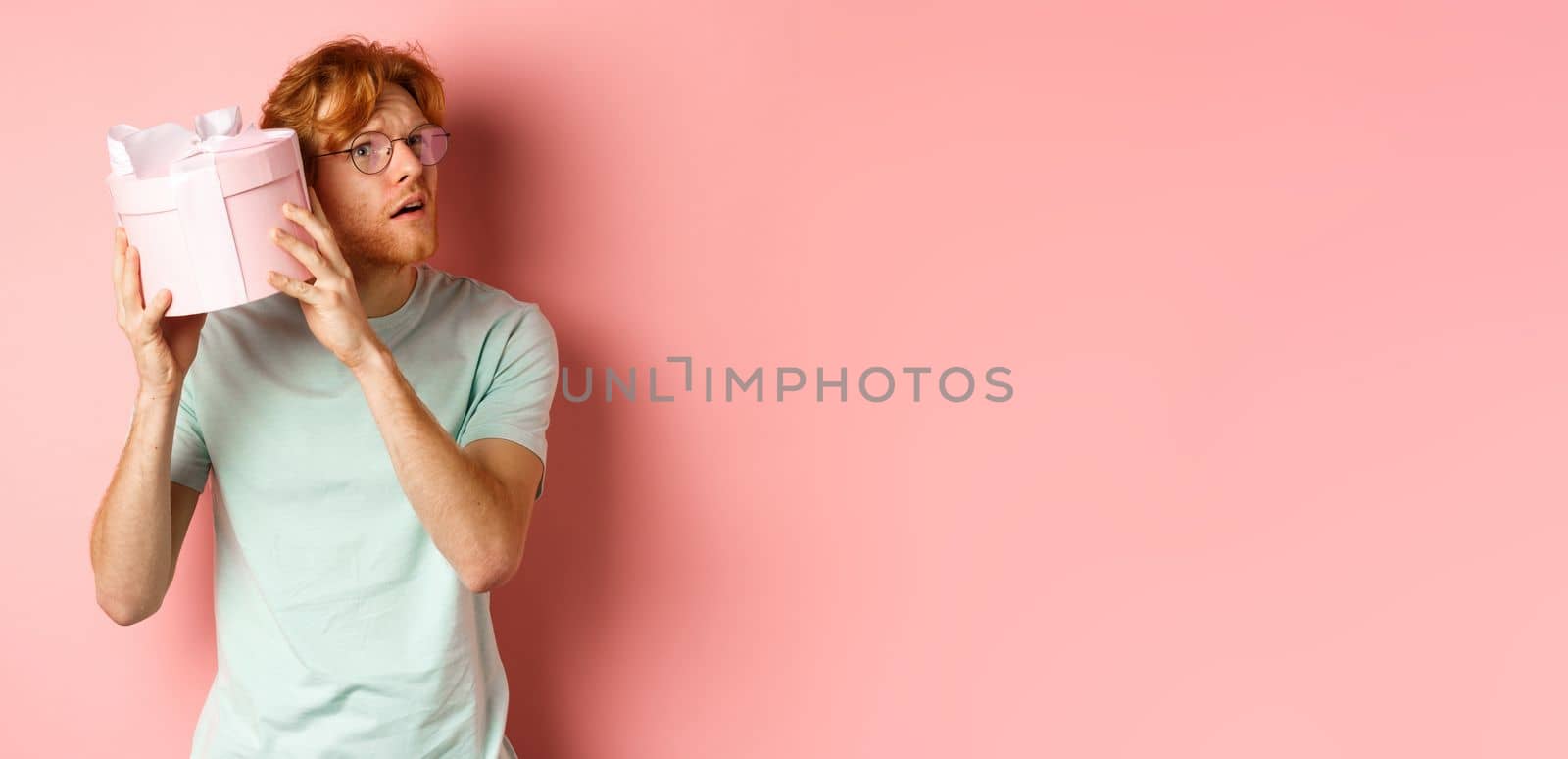 Valentines day and romance concept. Handsome redhead man shaking gift box and wonder what inside, trying to guess present, standing over pink background.