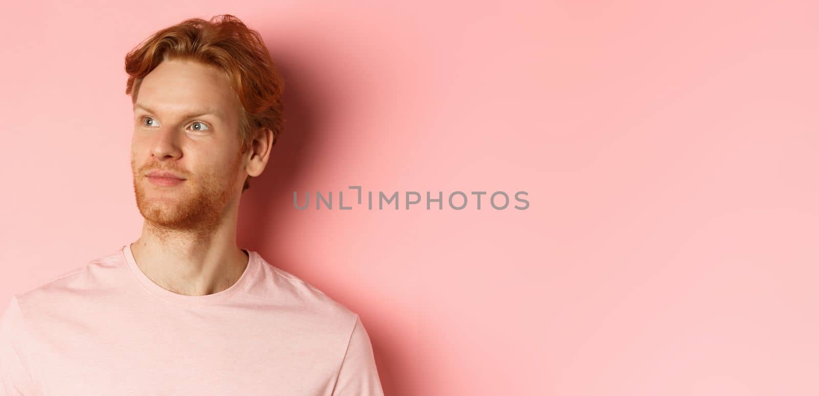 Handsome european male model with red hair and beard, turn head and looking pleased at copy space on left side, standing over pink background.