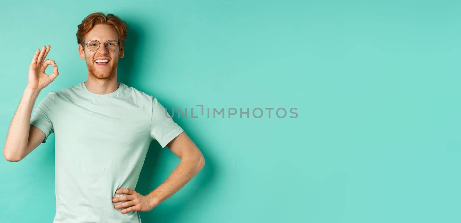 Happy young man with red hair and beard, wearing glasses and t-shirt, smiling satisfied and showing ok sign, say yes, approve and agree, standing over turquoise background by Benzoix