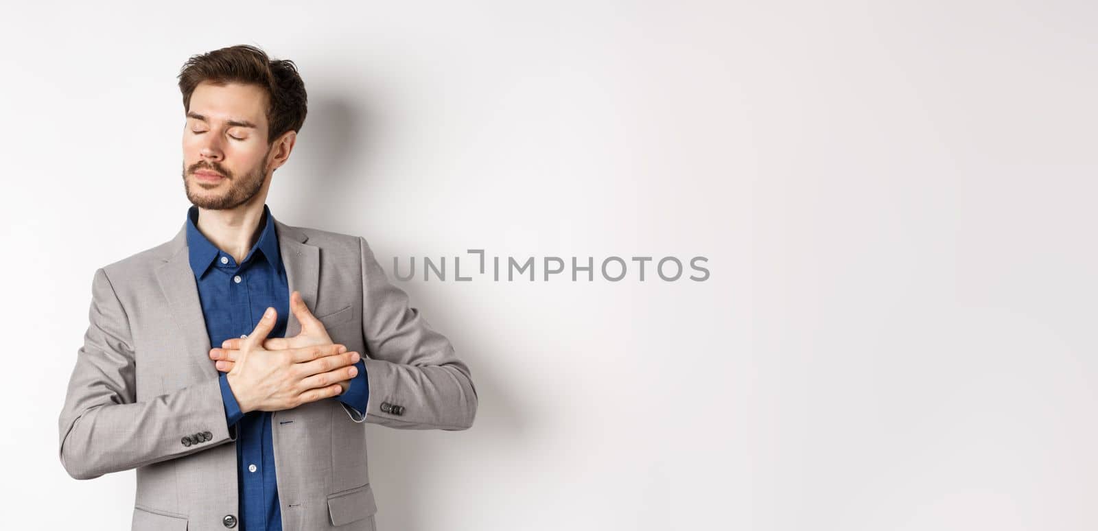 Romantic bearded man in stylish suit holding hands on heart, close eyes and remember something with tender feelings, standing nostalgic on white background.