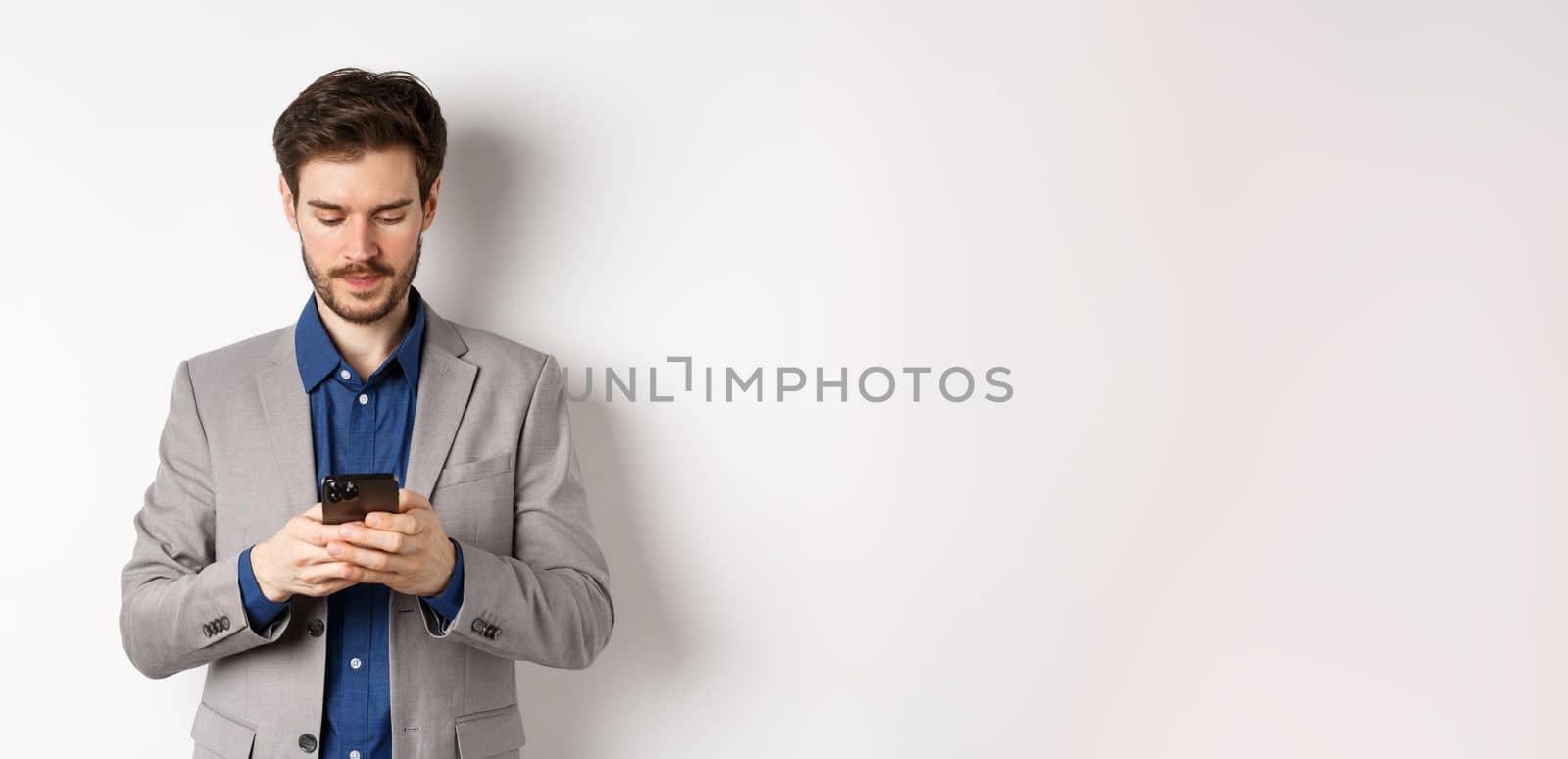 Businessman chatting on mobile phone, standing in suit with serious face, white background.