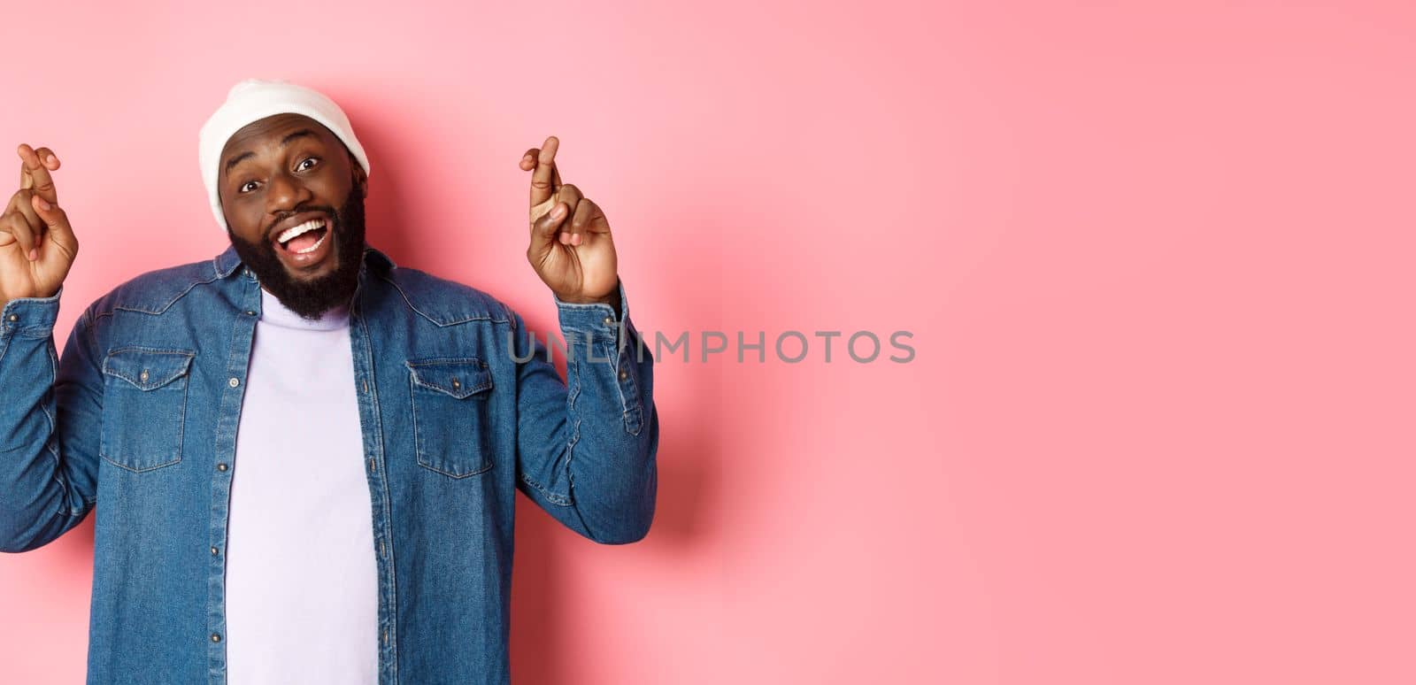 Optimistic african-american man making wish, holding fingers crossed and smiling, standing over pink background by Benzoix