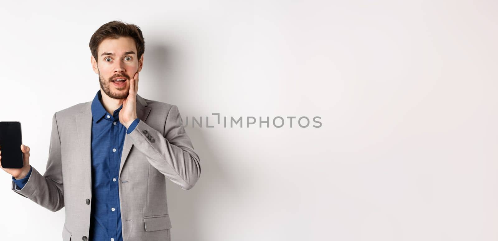 Excited business man in suit showing empty smartphone screen and gasping amazed, demonstrate application, standing on white background by Benzoix