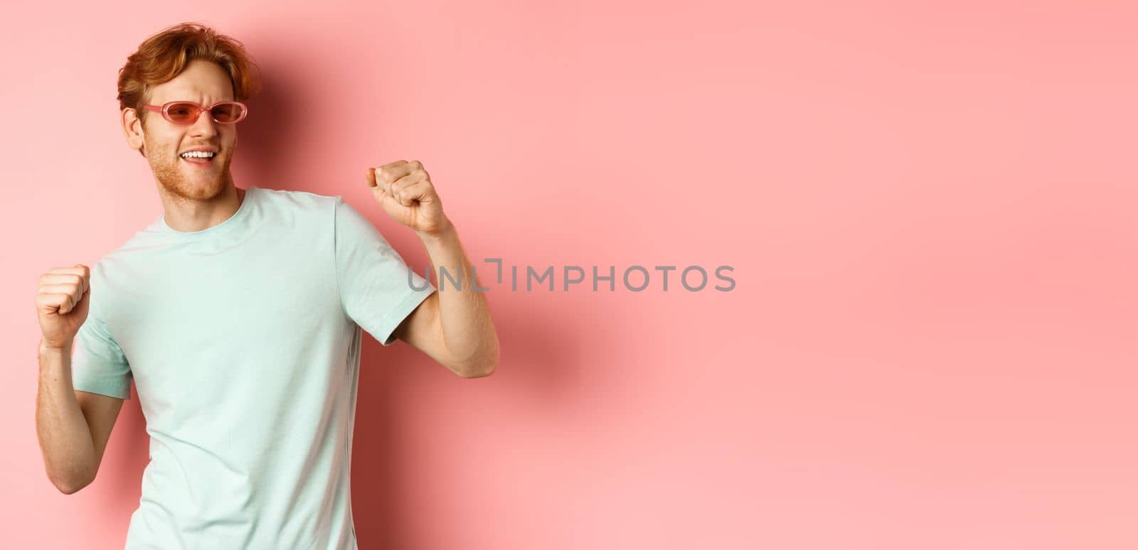 Tourism and vacation concept. Cheerful redhead man having fun at party, dancing and enjoying holiday, standing in sunglasses and t-shirt against pink background.