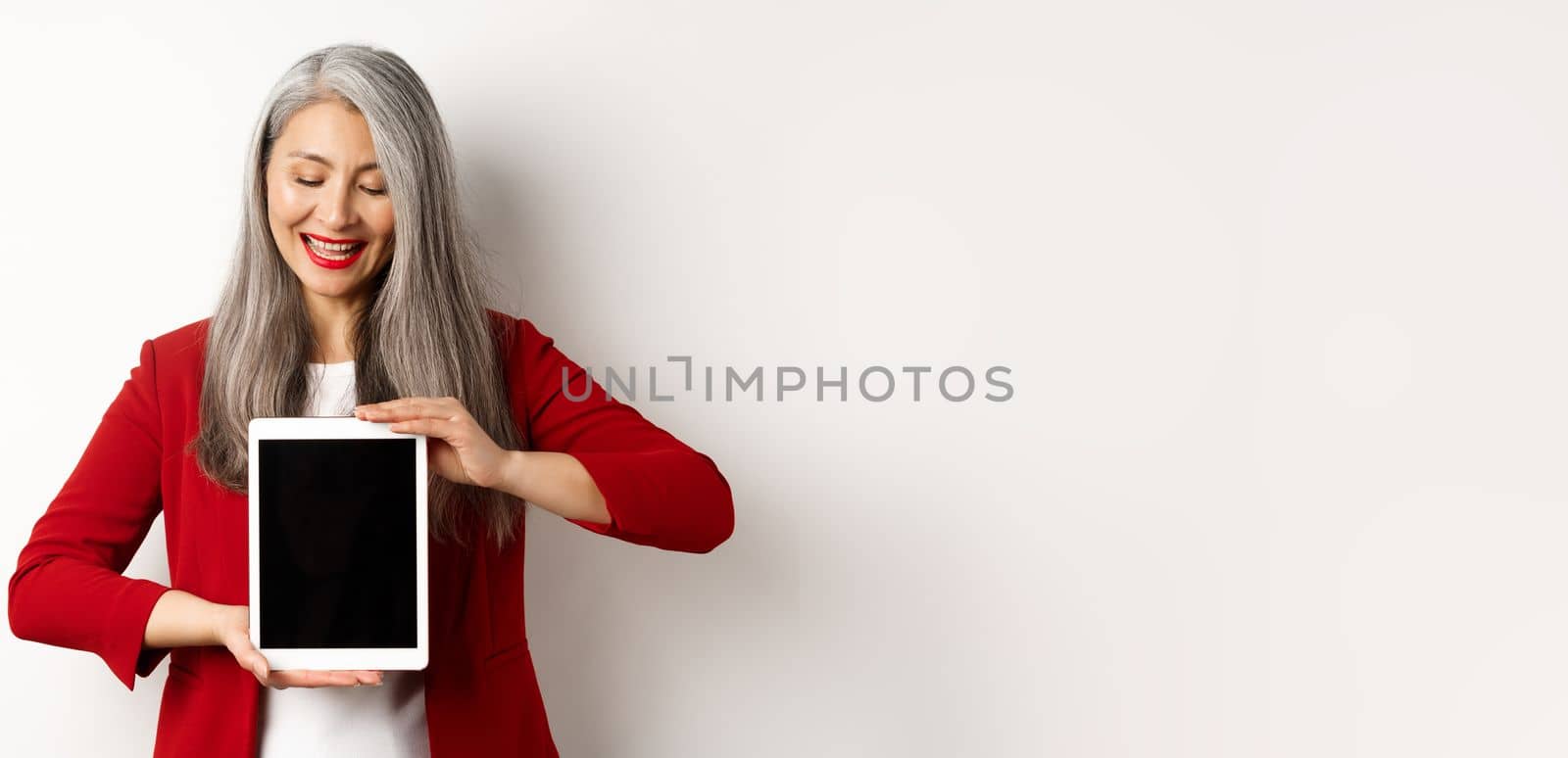 Business. Successful asian businesswoman in red blazer showing blank digital tablet screen, looking down with pleased smile, white background.